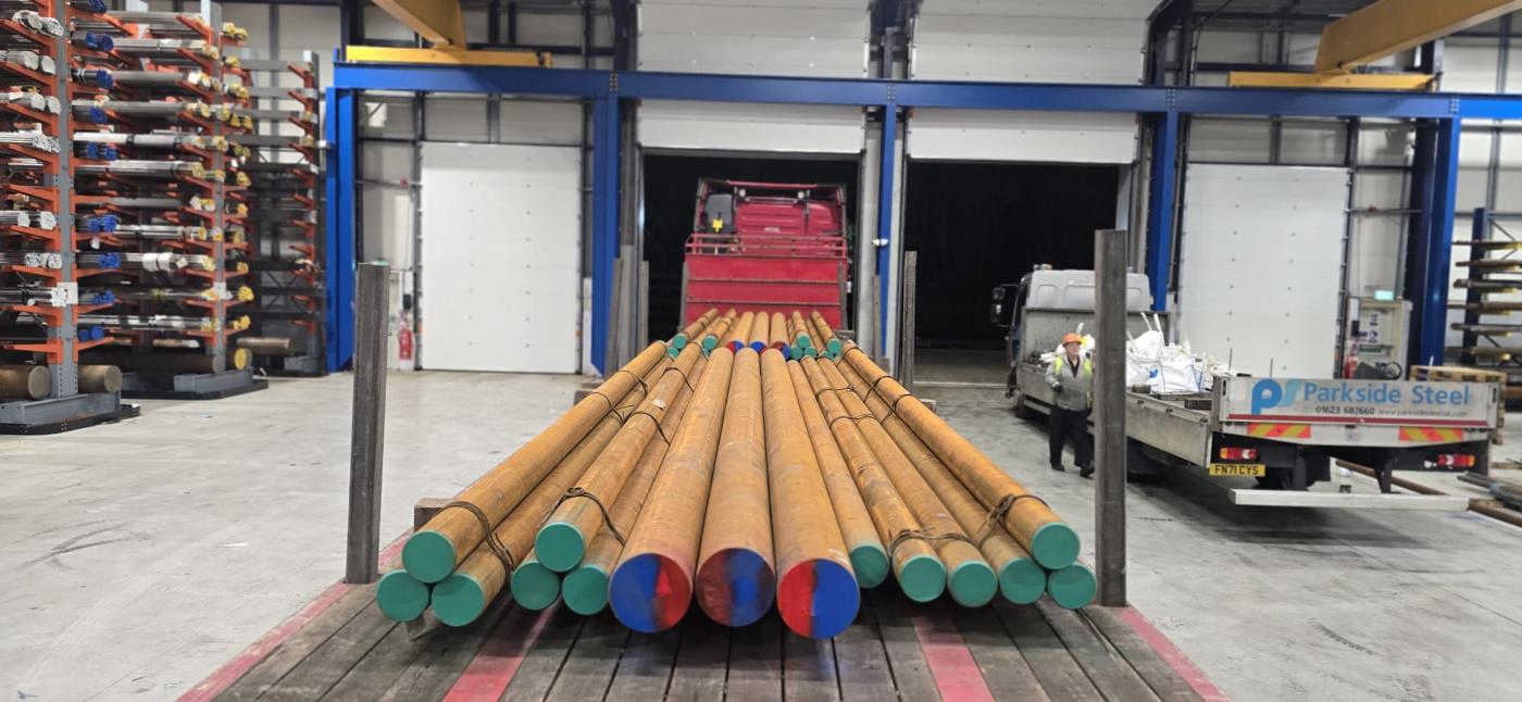 Large-diameter black round bar on a flatbed in the loading bay at Byron House, mill-coated with colour-coded ends, alongside a Parkside Steel delivery vehicle