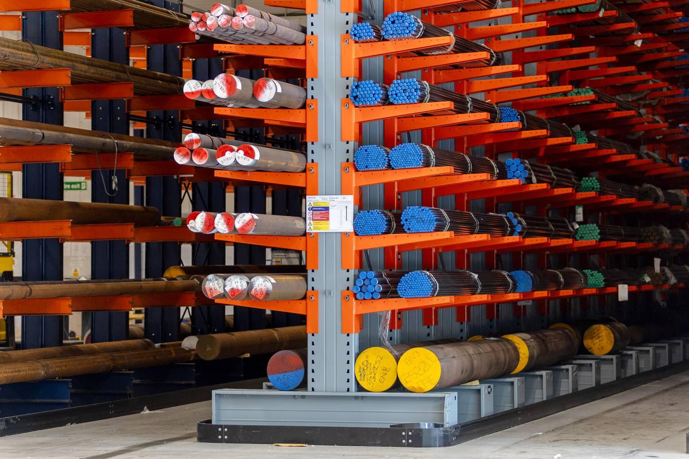 Orange cantilever racking showing red/white coded EN19 alloy bar alongside blue-coded bright bar — full range of engineering steel grades