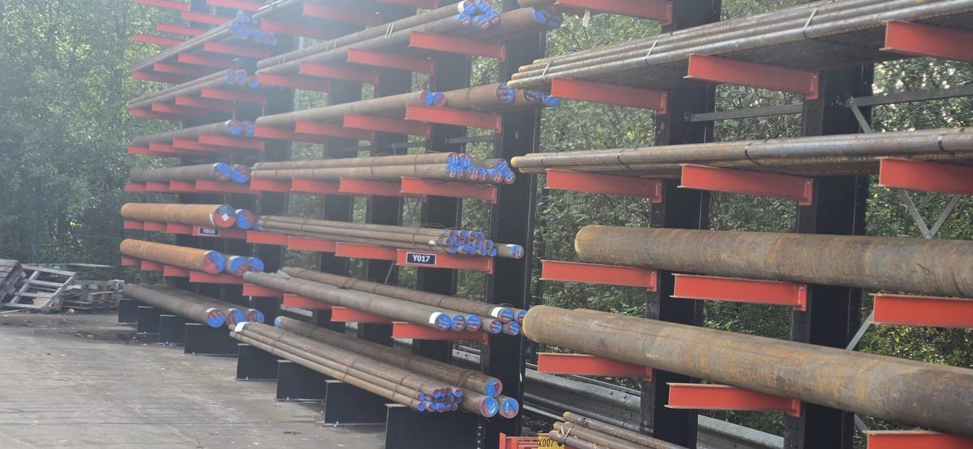Cantilever arms on the new external racking loaded with black round bar, bar ends paint-marked and each level carrying a rack location label