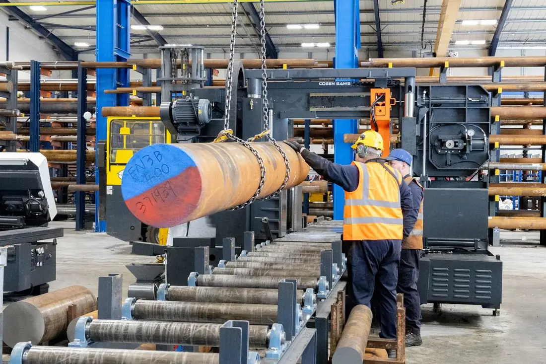 Two Parkside Steel operators loading a large EN3B black round bar onto the COSEN bandsaw using overhead chain crane